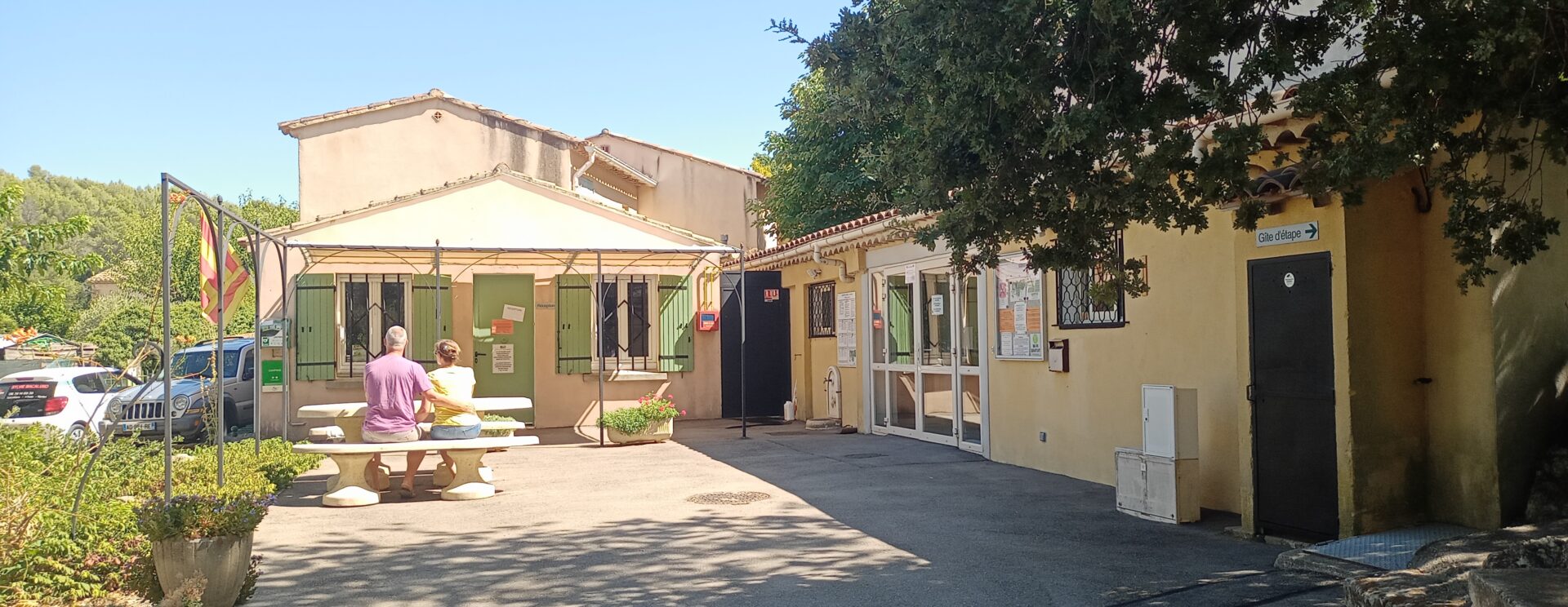 Vue de l'entrée du camping Les Royères du Prieuré de Maubec (accueil et sanitaire) avec en fond. Un couple est assis à la table