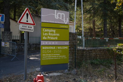 Vue du totem d'entrée du camping gris et vert (logo, nom du camping, pictogrammes de services, horaires) - Agrandir l'image, fenêtre modale